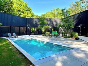 Une piscine rectangulaire à l’eau d’un bleu limpide, entourée d’une terrasse dallée. On y trouve des chaises longues et une petite table avec une chaise.