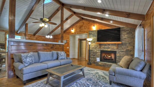 A spacious living room with a stone fireplace, wooden beams, and a ceiling fan. - Large Cabin Near the town of Blue Ridge! (Blue Ridge)