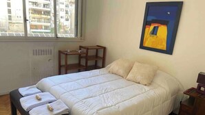 Une chambre à coucher comprenant un lit, un banc, une petite table, un radiateur et une fenêtre donnant sur des immeubles.