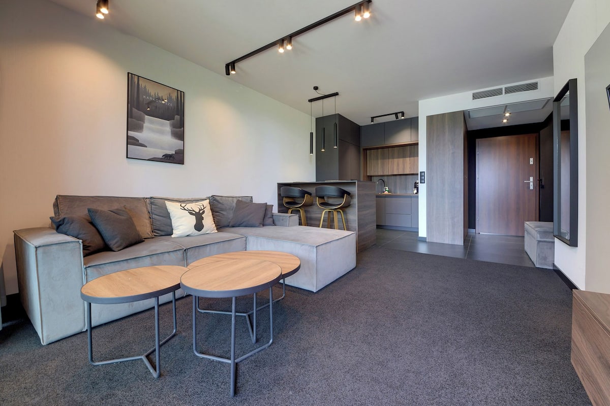 A modern living room with a grey sofa, two wooden tables, and a kitchen in the background.
