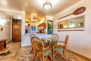 A dining area with a round glass table and wicker chairs, a wooden door, and a kitchen in the background. - Menehune Shores, #220^ 2 Bedroom Condo by RedAwning (Kihei)