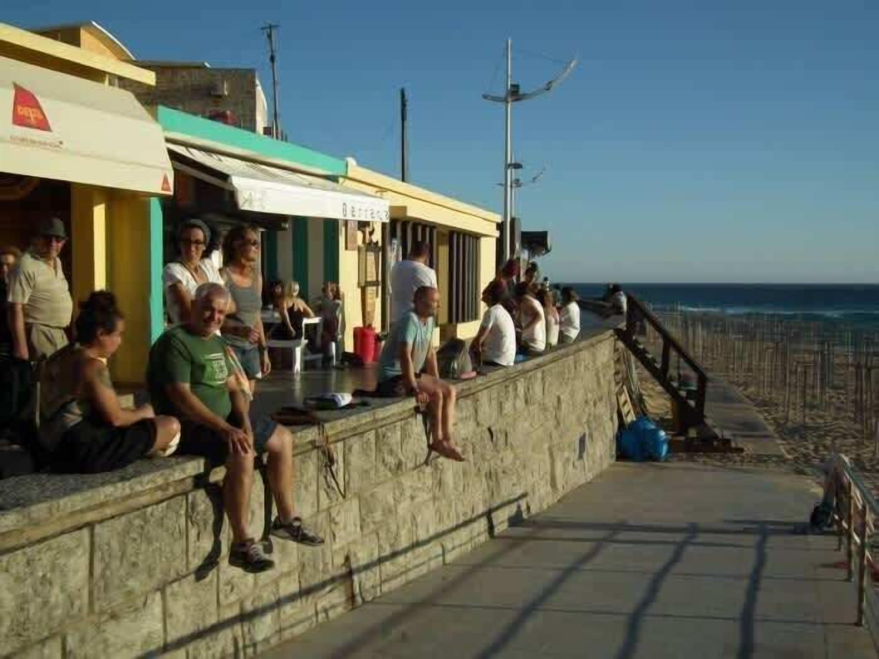 Une promenade en bord de mer, avec des personnes assises sur un muret de pierre, d’où l’on a une vue sur la plage.