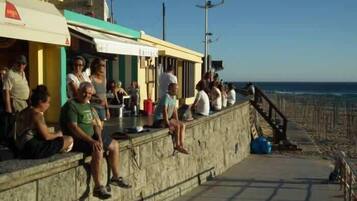 Une promenade en bord de mer, avec des personnes assises sur un muret de pierre, d’où l’on a une vue sur la plage.