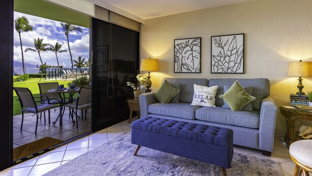 A living room with a grey sofa, a blue ottoman, and a view of a garden with palm trees.