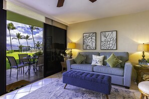 A living room with a grey sofa, a blue ottoman, and a view of a garden with palm trees. - Kihei Surfside #102 1 Bedroom Condo by RedAwning (Kihei)