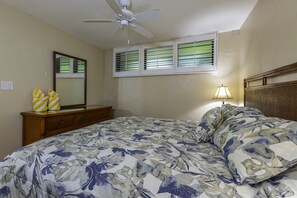 A bedroom with a bed, a ceiling fan, a dresser, a mirror, and a window with blinds. - Kihei Surfside #102 1 Bedroom Condo by RedAwning (Kihei)