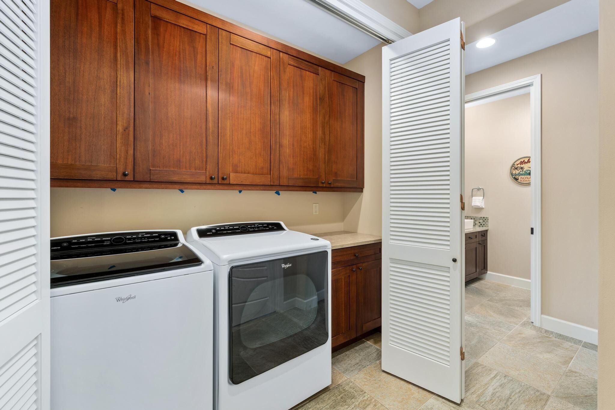 A laundry room with a Whirlpool washer and dryer, wooden cabinets, a white door, and a tiled floor.