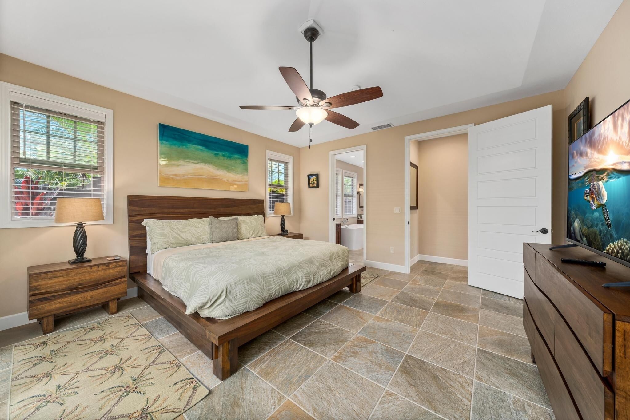 Une chambre à coucher avec un lit, des tables de chevet, un ventilateur de plafond, un tableau, un téléviseur et une salle de bain visible à travers une porte ouverte.