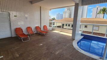 A poolside area with three orange lounge chairs, a tiled wall, and a view of tall buildings.