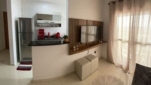 A modern kitchen with a refrigerator, stove, and microwave. There is a wooden cabinet and a television mounted on the wall. A small seating area with a round table and a cushion is visible. - Excelente Apto com piscina em Praia Grande (Praia Grande)