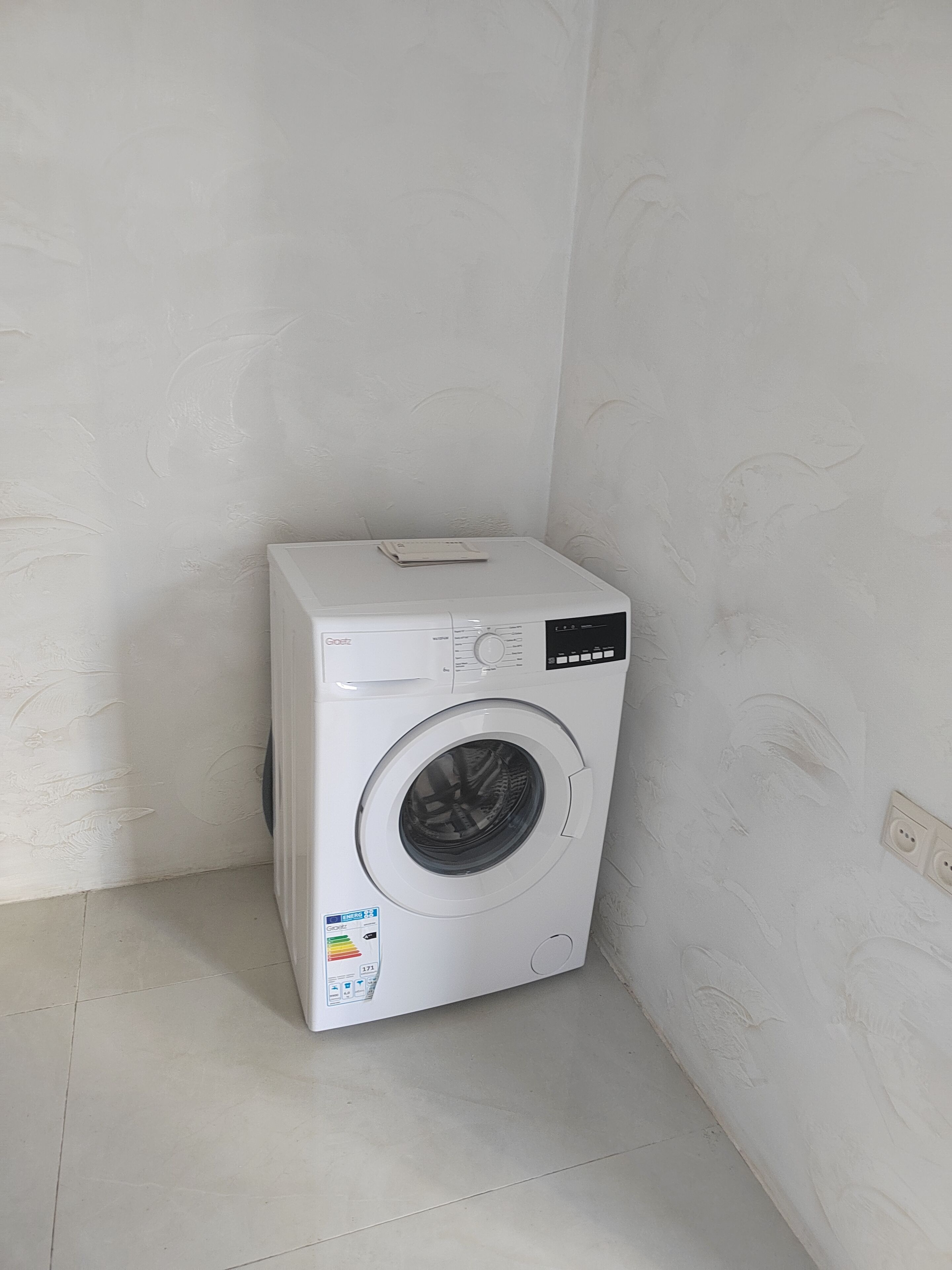 A compact, white washing machine in a corner of a room with plain walls and a tiled floor.