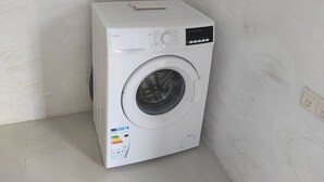 Eine kompakte, weiße Waschmaschine steht in einer Ecke des Raumes mit schlichten Wänden und Fliesenboden.