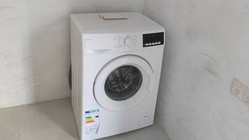 A compact, white washing machine in a corner of a room with plain walls and a tiled floor.