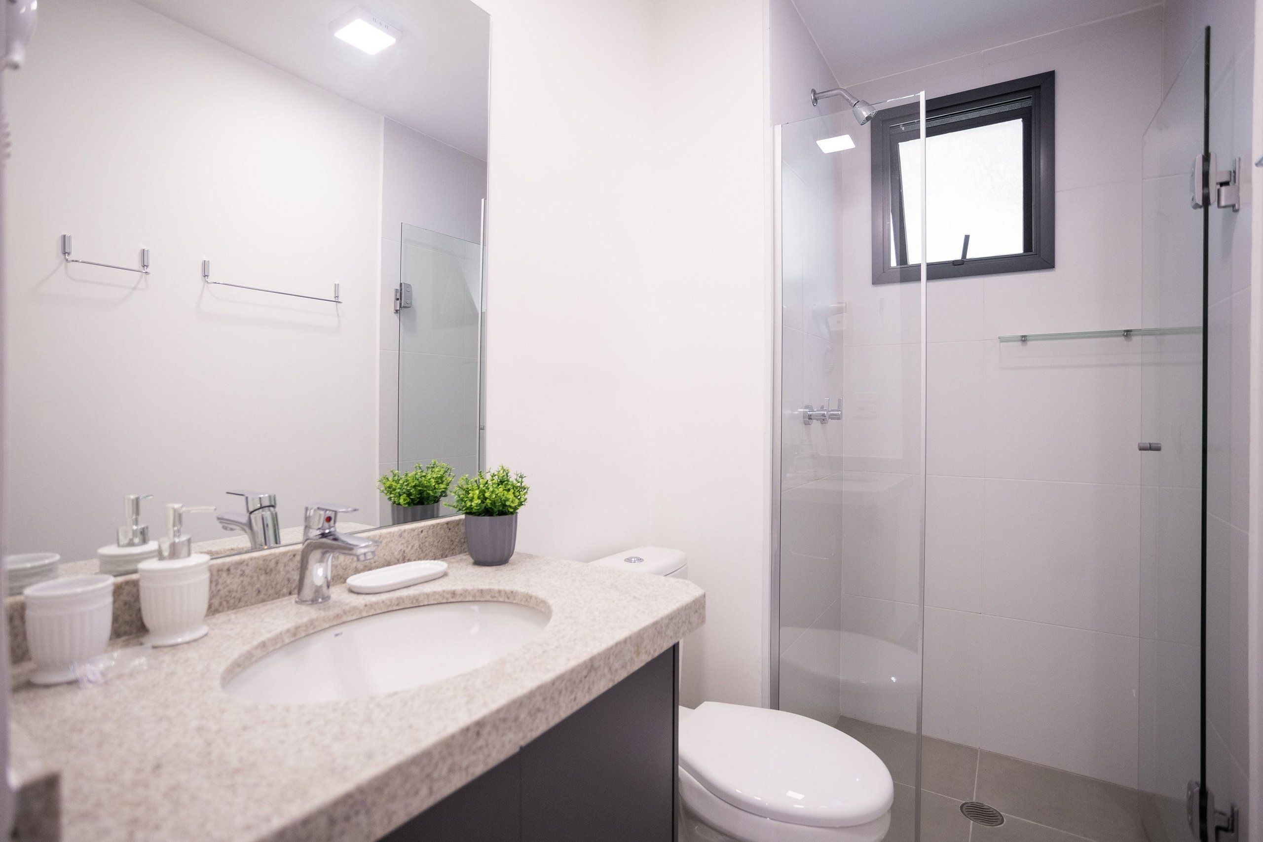 A modern bathroom with a granite countertop, white fixtures, a glass-enclosed shower, and a small window.