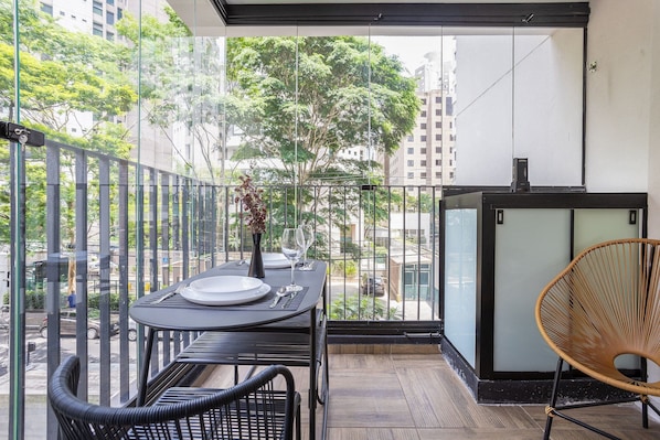 Uma varanda com uma mesa posta para dois, uma cadeira e vista para um horizonte urbano com árvores e edifícios.