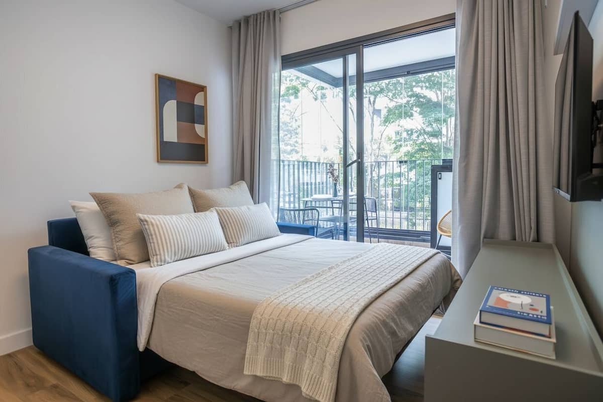 Ein Schlafzimmer mit einem Bett, einem blauen Sessel, einem kleinen Tisch und einem Balkon mit Blick auf Bäume.