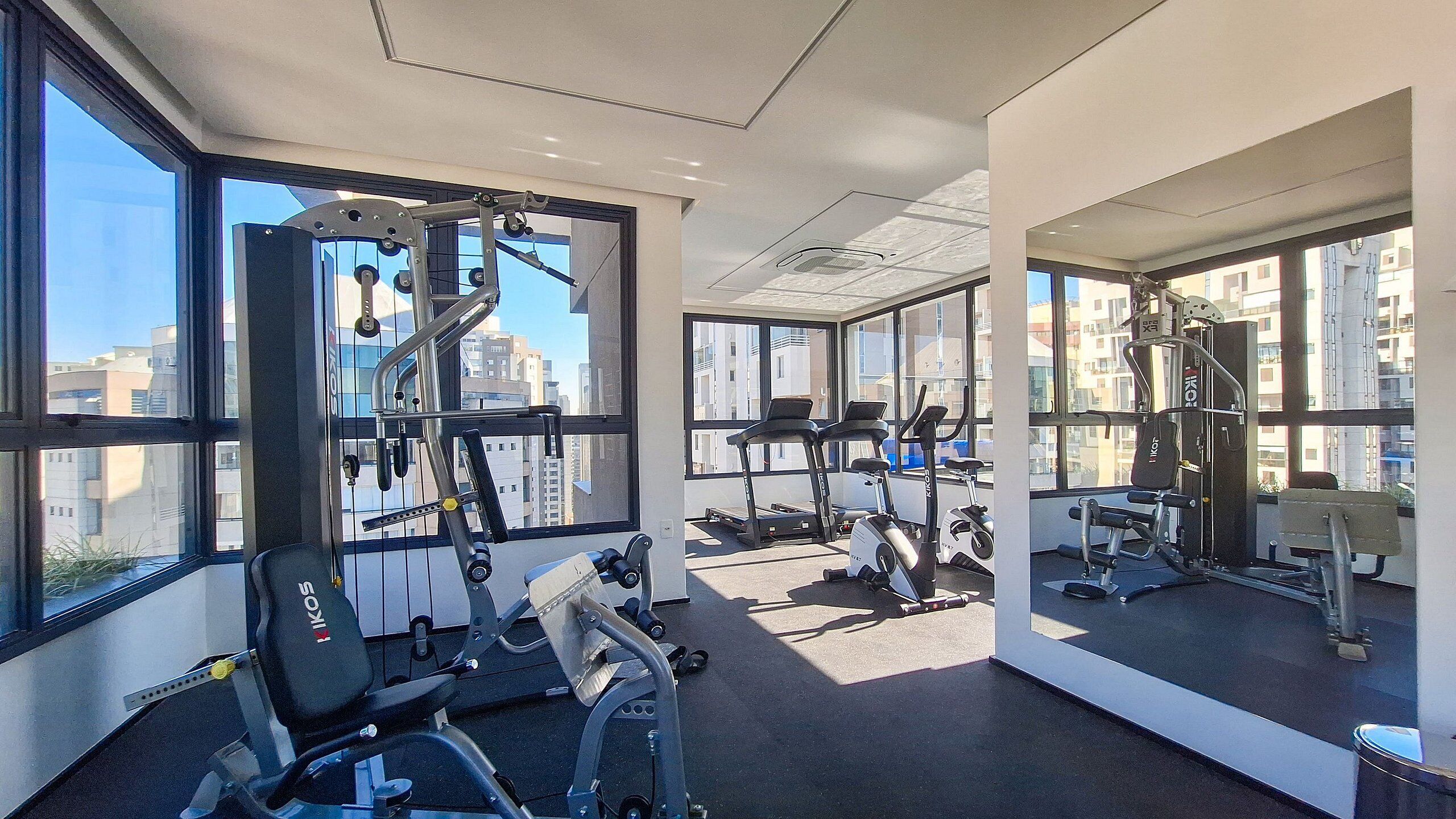 Uma academia moderna com diversos equipamentos de exercício, amplas janelas com vista para a cidade e um interior limpo e bem iluminado.