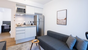 Una cocina moderna con armarios blancos, un microondas y un refrigerador. Hay un sofá gris con cojines y un taburete de madera.