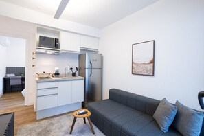 A modern kitchen with white cabinets, a microwave, and a refrigerator. There is a gray sofa with cushions and a small wooden stool. - Walk Moema (São Paulo)