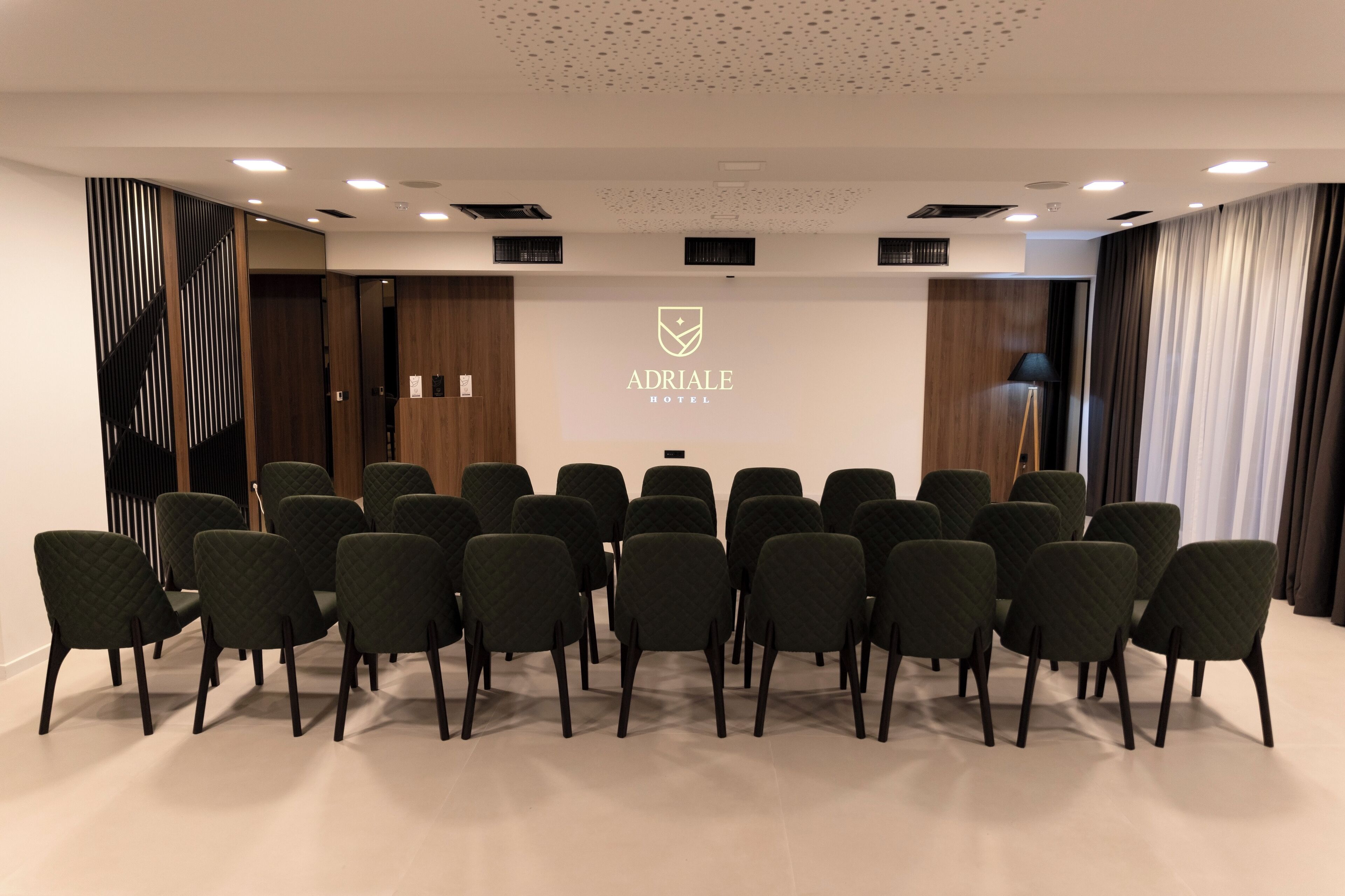 Una sala de conferencias con el logo de Adria Hotel, que presenta un diseño moderno con techo blanco, iluminación empotrada y una fila de sillas verde oscuro.