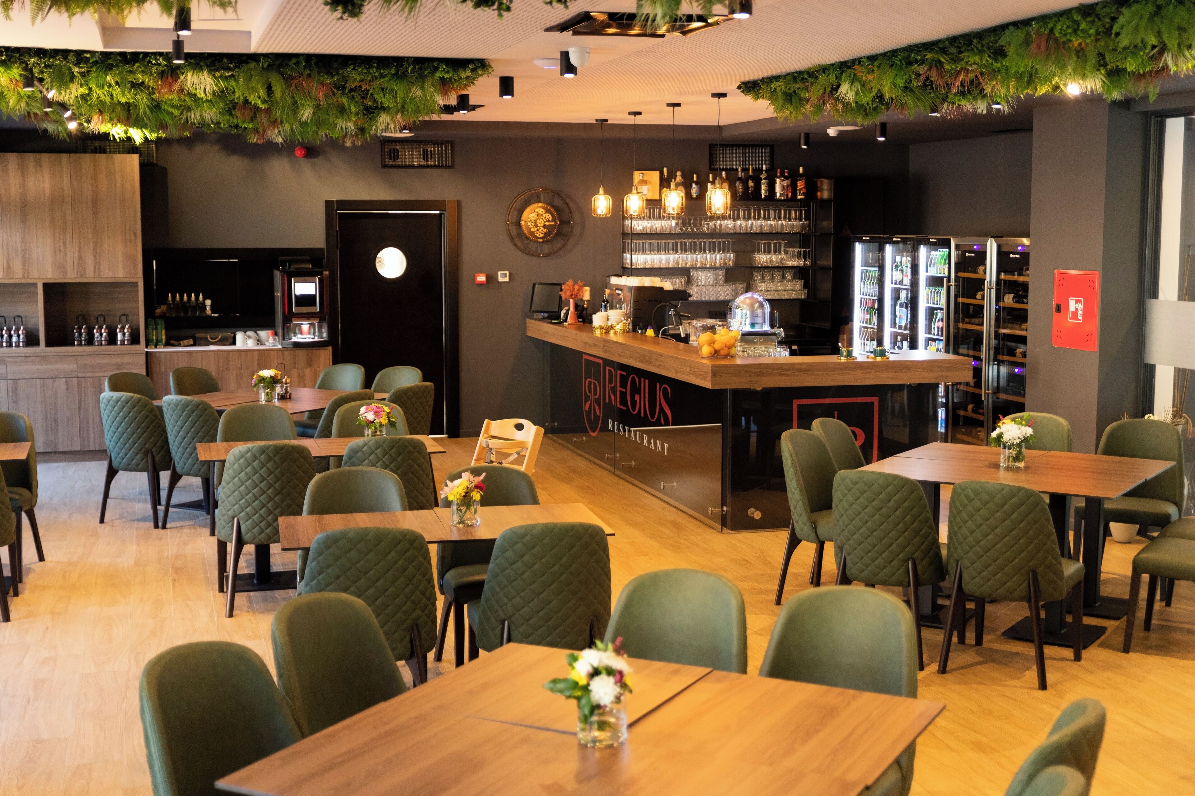 Een restaurant met groen bekleden stoelen, houten tafels, een bar en een klok aan de muur.