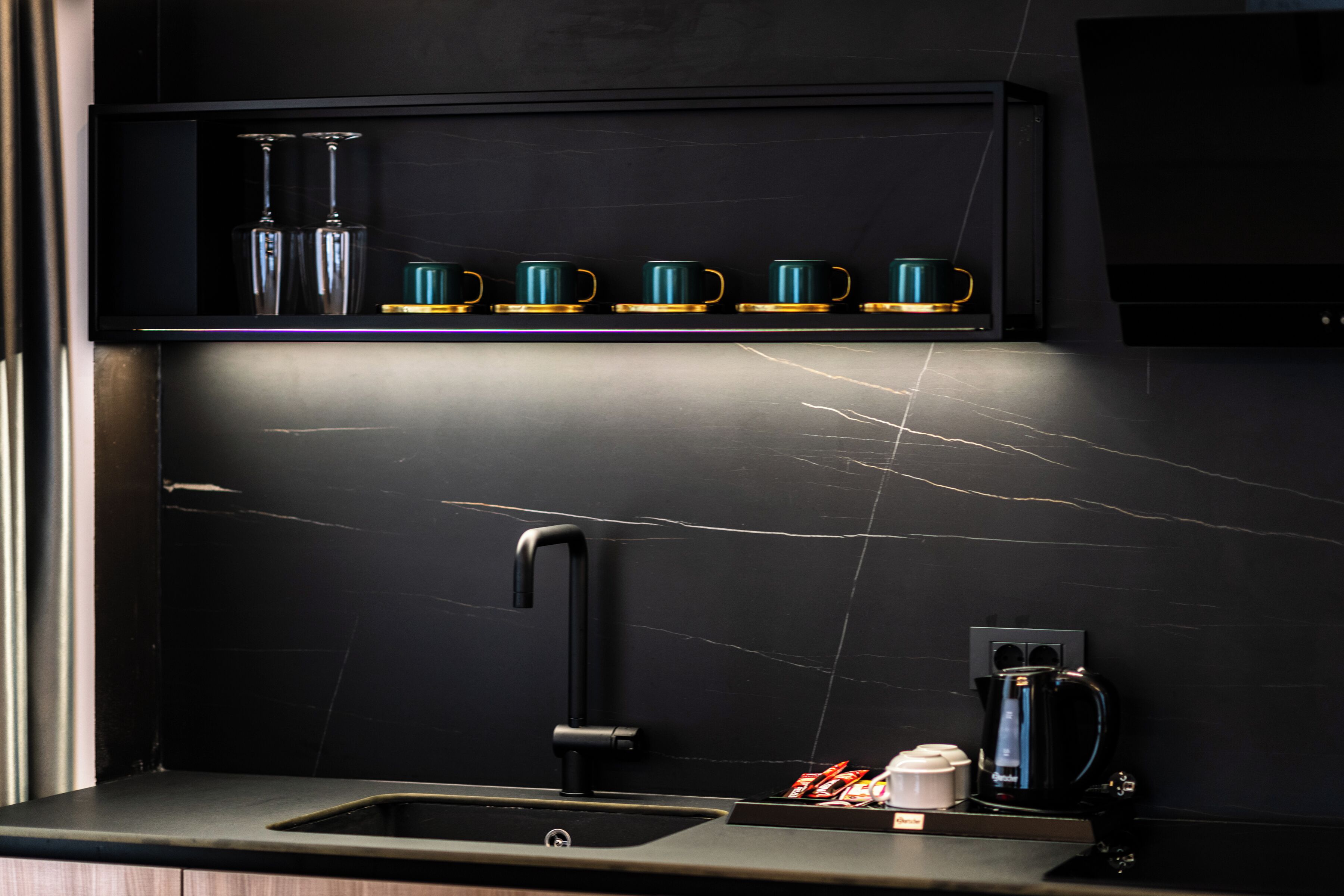 A modern kitchen with a black countertop, a single faucet, and a shelf above holding four teal mugs and two clear bottles.