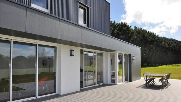 Une maison moderne dotée de grandes portes vitrées, d’une terrasse en bois et d’un coin repas.