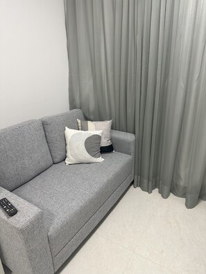 A grey sofa with a remote control on it, positioned against a grey curtain.