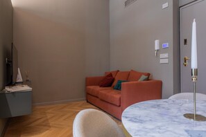Egy modern nappali, amelyben van egy kanapé, egy kerek asztal és egy televízió, mely egy állványon található.