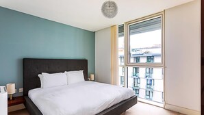 A modern bedroom with a large bed, a bedside table, a lamp, and a view of the city through a large window. - City-Close 1-Bed Apartment near Tower Hill (London)