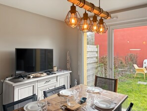 A dining area with a TV, wooden table, chairs, and a glass door leading to a garden.