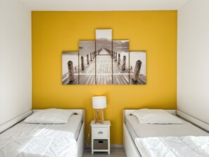A hotel room with two beds, a yellow wall, and a large artwork depicting a wooden pier.