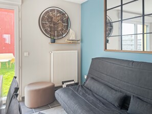 A modern living room with a grey sofa, a round wall clock, a radiator, and a window with blinds.