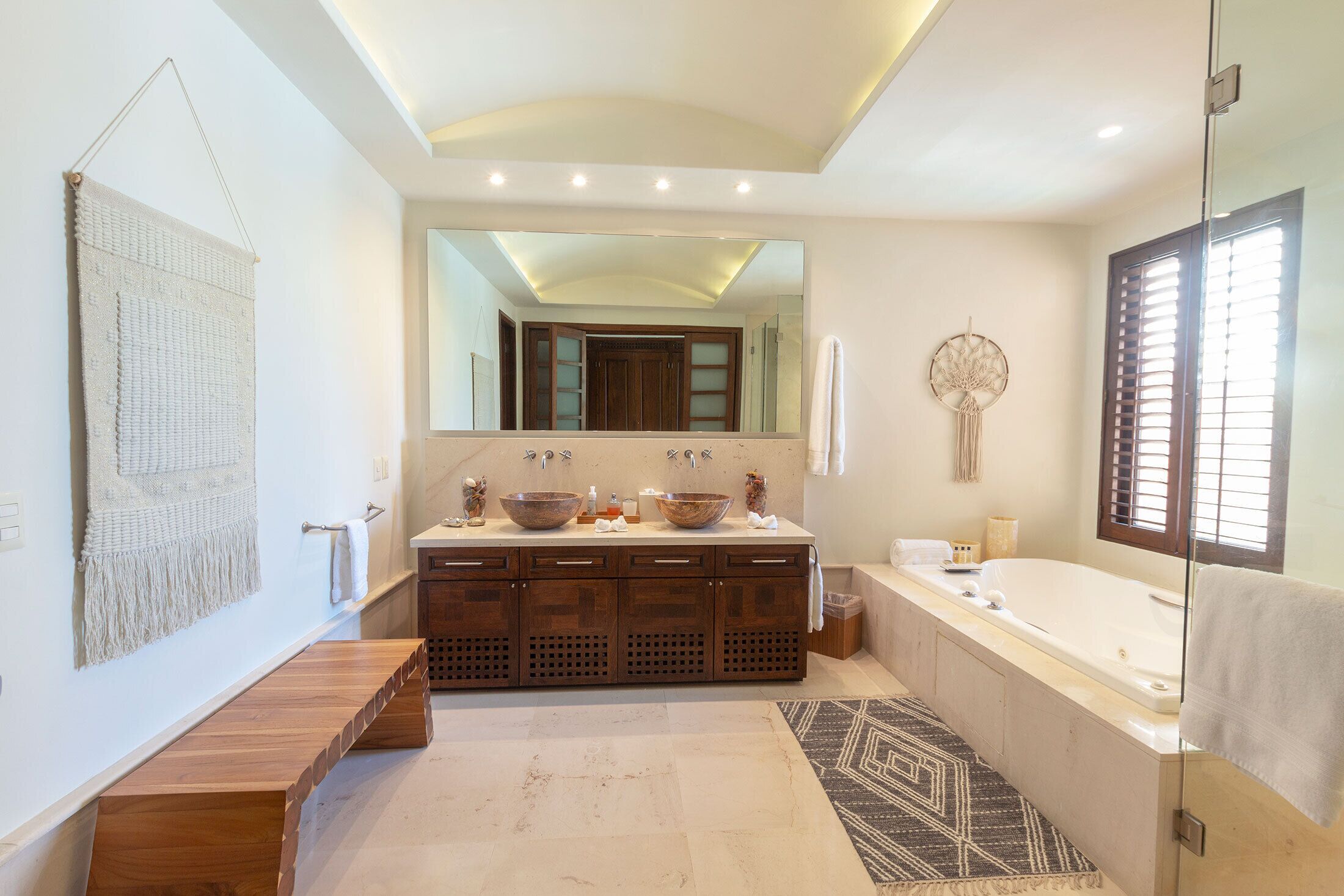 A modern bathroom with a large mirror, double sinks, a bathtub, and a wooden bench.