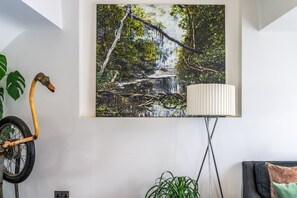 Una habitación con un cuadro colgado en la pared que muestra un arroyo en un bosque, una lámpara de pie, una bicicleta y una planta en maceta.