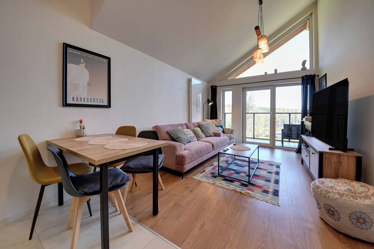A modern living room with a wooden dining table, a sofa, a TV, and a large window.