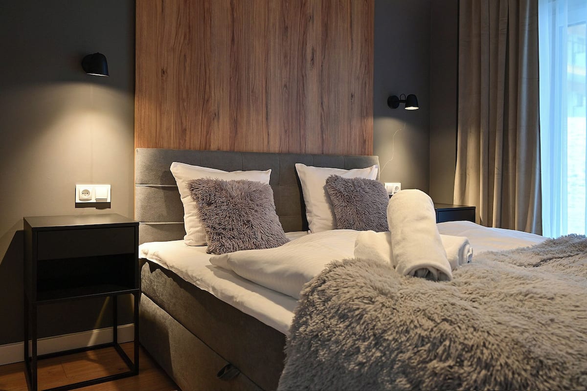 A modern bedroom with a wooden headboard, two fluffy pillows, a neatly made bed, and a nightstand with a lamp.