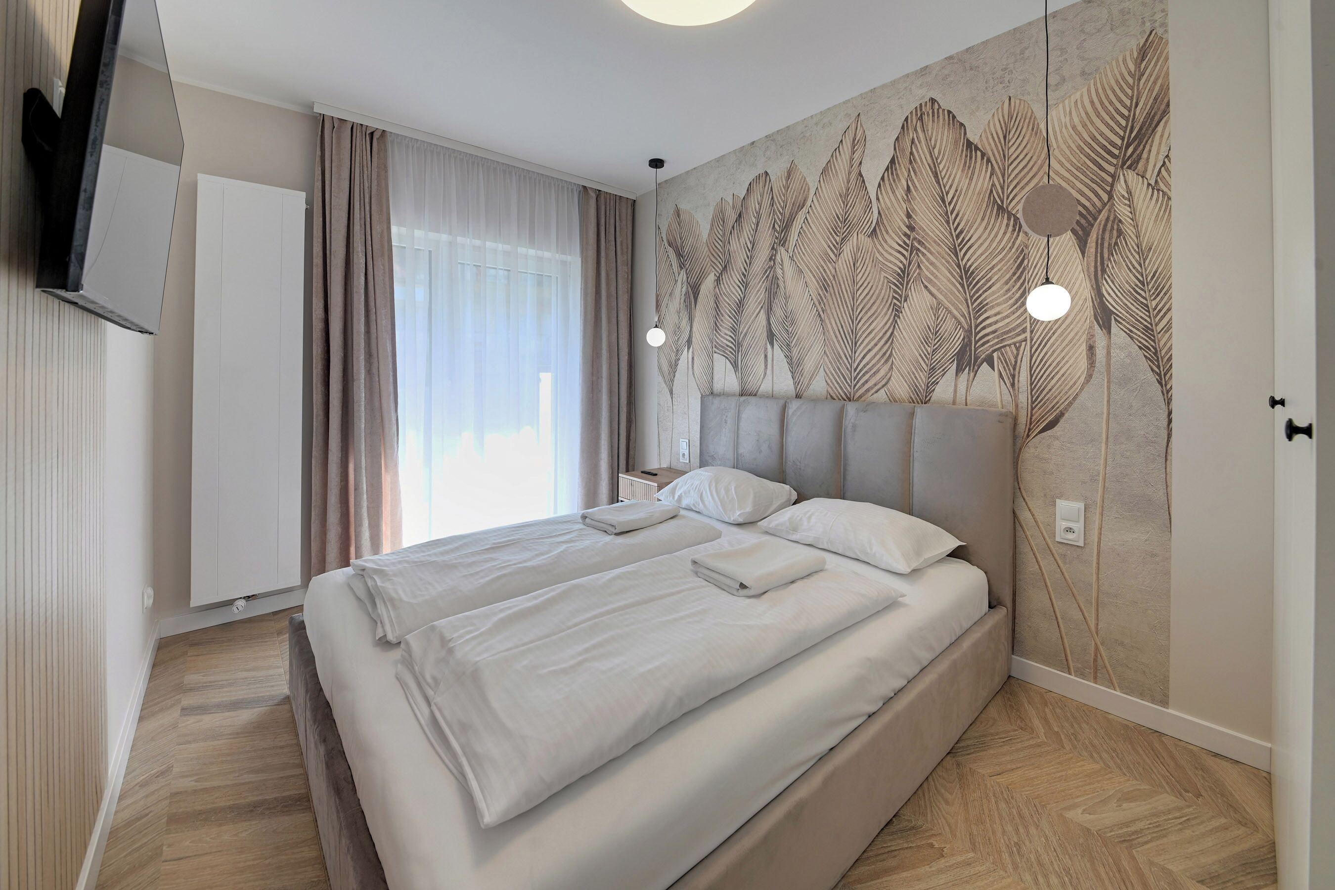 Ett modernt hotellrum med en stor säng, en platt-TV, en väggmonterad lampa och ett stort fönster med gardiner.