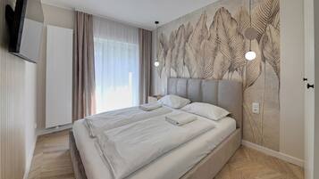 Ett modernt hotellrum med en stor säng, en platt-TV, en väggmonterad lampa och ett stort fönster med gardiner.