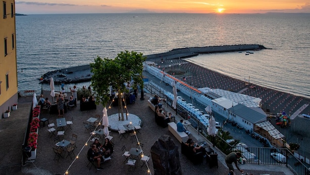 A seaside terrace with outdoor seating, a view of the sea, and a building with a balcony.