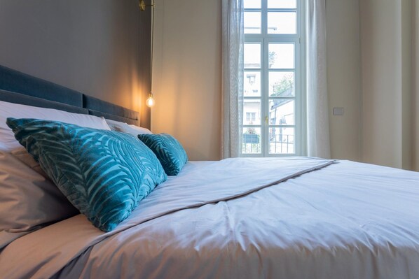 A neatly made bed with white linens and two teal pillows, a bedside lamp, and a window with curtains. - Lagrange Design Apartments - Beta by Wonderful Italy (Turin)