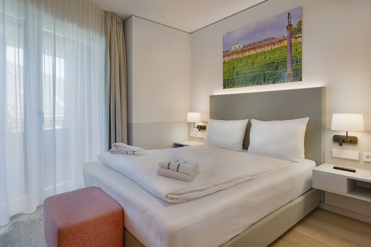 A neatly made bed with white linens and two pillows, a bedside table with a lamp, a window with sheer curtains, and a painting of a building on the wall.