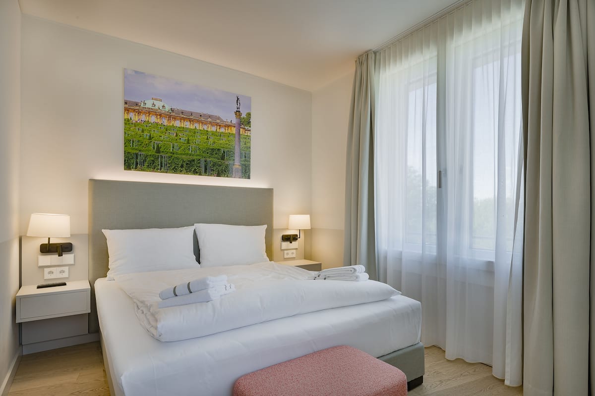 A hotel room with a neatly made bed, two bedside tables with lamps, a painting on the wall, and a large window with curtains.