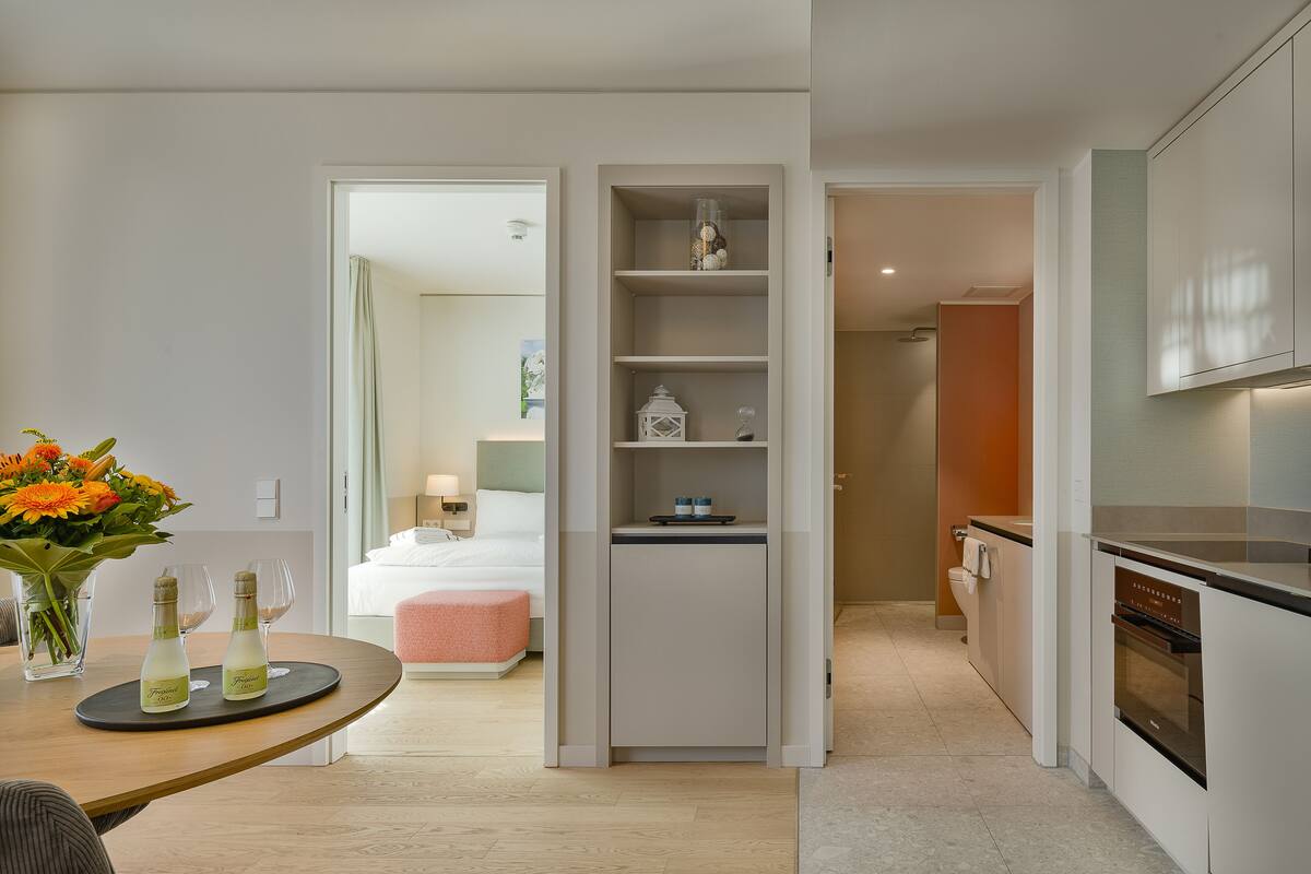 A modern hotel room with a kitchenette, a dining area with a table and chairs, a bed with a pink ottoman, and a bathroom visible through an open door.
