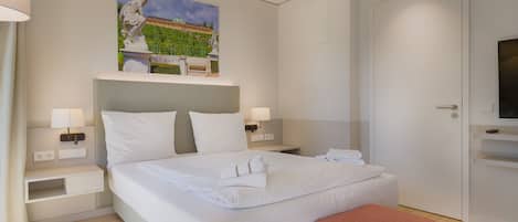 Una habitación de hotel moderna con una cama bien hecha, un cuadro sobre la cama, un televisor en un mueble bajo y una puerta a la derecha.