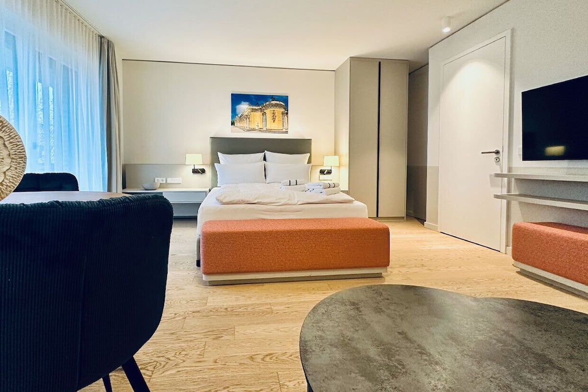 A modern hotel room with a bed, bedside tables, a TV, and a large window with curtains.