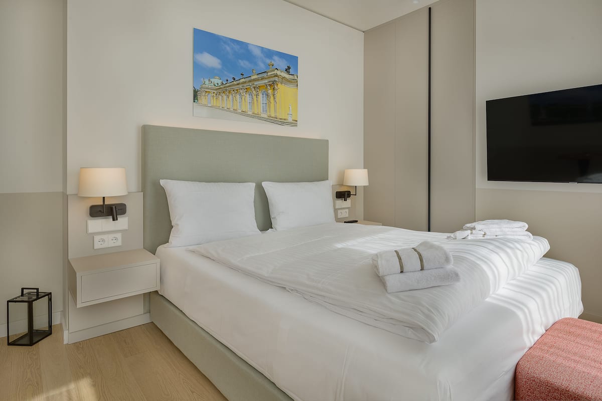 A neatly made bed with white linens and pillows, a bedside table with a lamp, a small bedside table with a book, and a painting of a building on the wall.