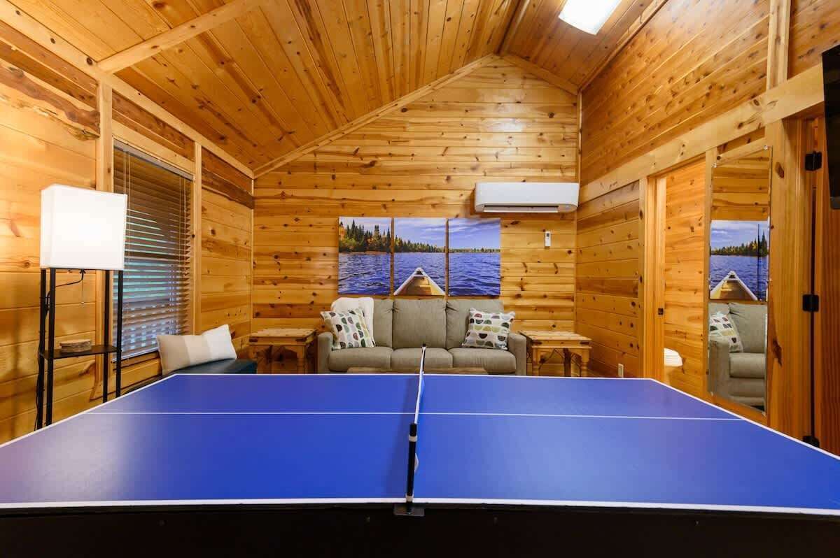 Interior de una cabaña de madera con mesa de ping pong, sofá y ventana con persianas.