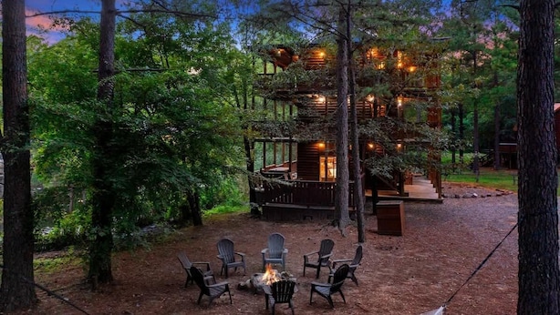 En hytte omgivet af træer med et bålsted og stole i forgrunden.