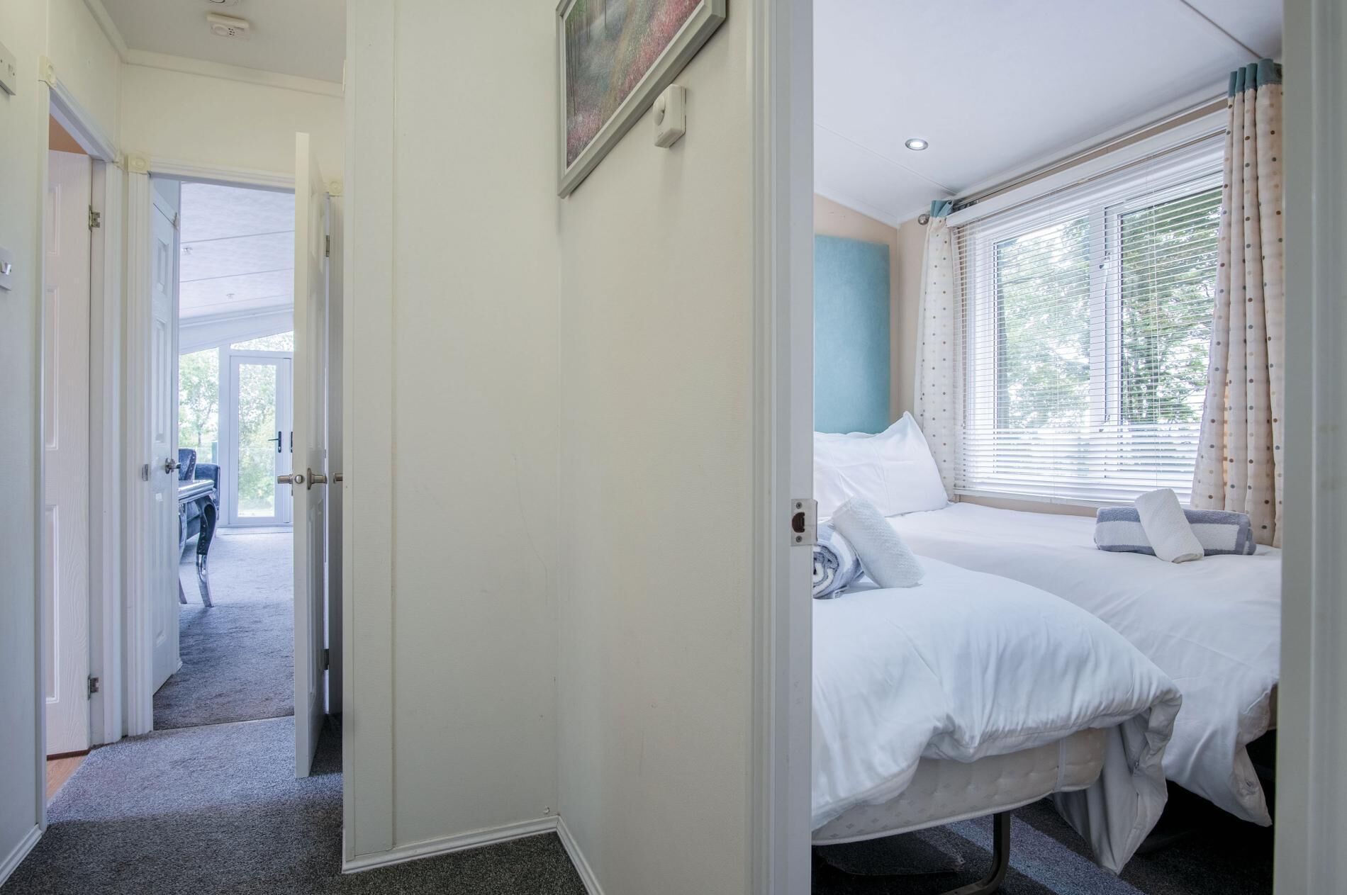 Une chambre d’hôtel avec deux lits, une fenêtre avec des stores, une porte donnant sur un couloir et un tableau accroché au mur.